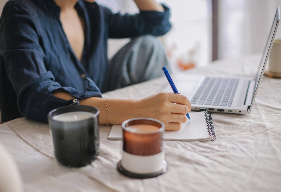 woman at a table with a laptop and notepad in front of her. She is writing with pen on the notepad